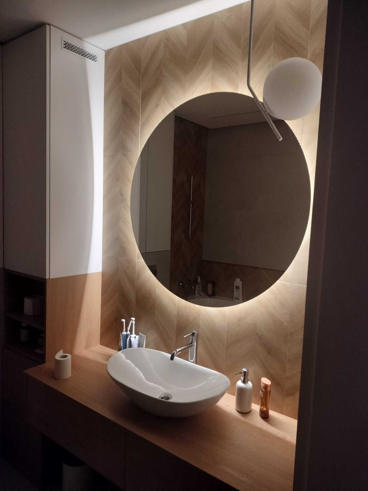 Nowoczesna łazienka z okrągłym lustrem podświetlanym LED, umywalką nablatową, drewnianym blatem i płytkami ułożonymi w jodełkę na ścianie.