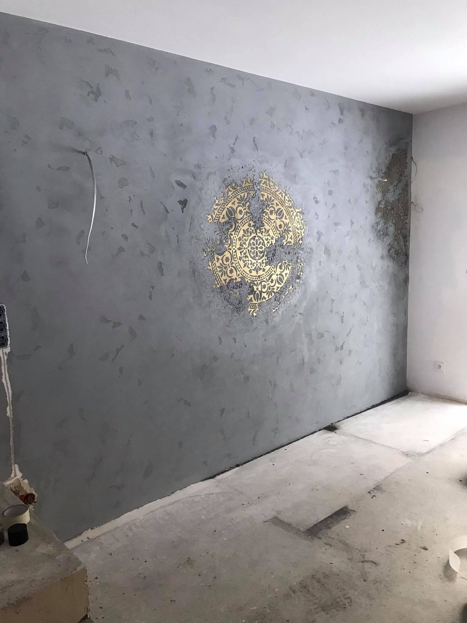 Szara ściana z dekoracyjną, złotą rozetą w stylu mandali, widoczne niedociągnięcia wykończeniowe i wystające kable.