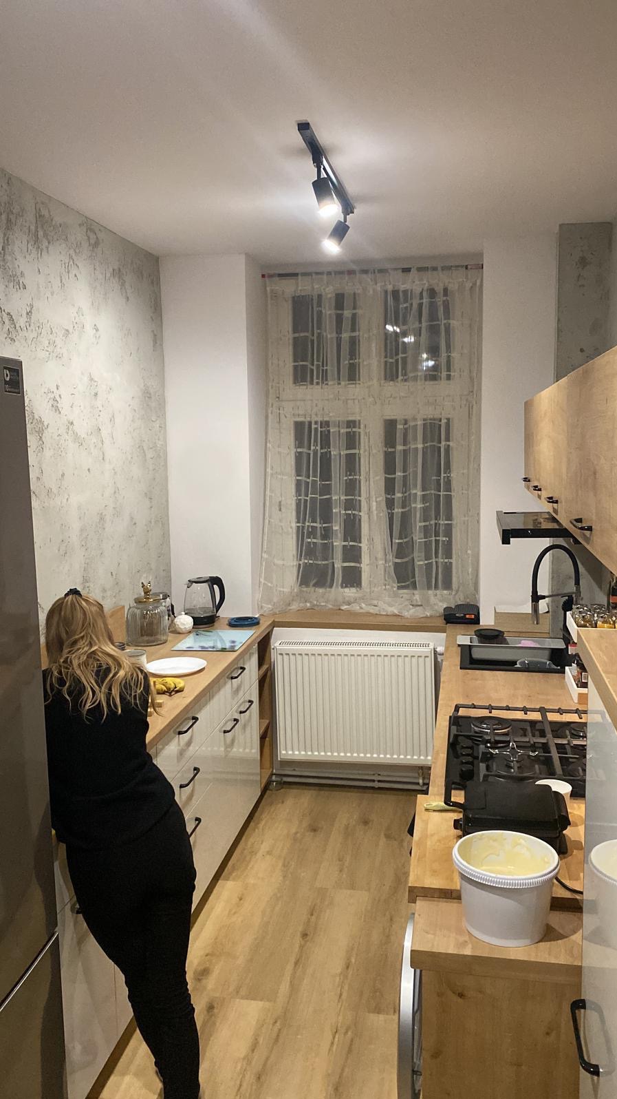 Wnętrze kuchni po remoncie: jasne szafki z drewnianymi blatami, ściana z dekoracyjnym tynkiem, okno z firanką, nowoczesne oświetlenie punktowe i kobieta przygotowująca posiłek.