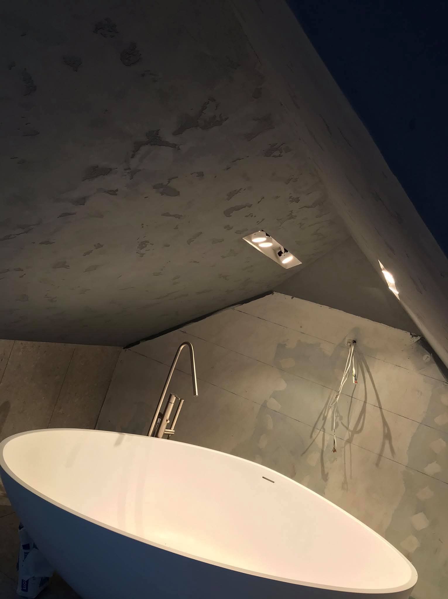 Wanna wolnostojąca z baterią w surowym wnętrzu łazienki, widoczne niedokończone ściany i wystające przewody elektryczne, oświetlenie punktowe w suficie.