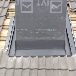 Ekspert-Dach Pogotowie Dekarskie - Szczegółowe ujęcie komina dachowego z obróbką blacharską i membraną izolacyjną Aqua Control XL, na tle dachówki ceramicznej. Widoczne drewniane elementy konstrukcji dachu.