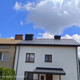 Ekspert-Dach Pogotowie Dekarskie - Biały dom z nowym, grafitowym dachem blaszanym na tle błękitnego nieba z chmurami. Widoczny fragment sąsiedniego budynku z jasnobrązowym dachem.