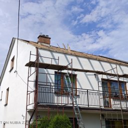 Ekspert-Dach Pogotowie Dekarskie - Dom w trakcie wymiany dachu, widoczne rusztowania i częściowo zdjęte pokrycie. Fragmenty starej więźby dachowej i komin z cegły. Błękitne niebo w tle.