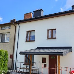 Ekspert-Dach Pogotowie Dekarskie - Biały dom z nową, ciemną obróbką blacharską dachu i rynnami. Widoczny fragment rusztowania i osoba w trakcie prac wykończeniowych przy wejściu.