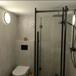 Nowoczesna łazienka z szarymi płytkami imitującymi beton, czarną kabiną prysznicową typu walk-in, podtynkową toaletą i okrągłymi lampami ściennymi z ciepłym światłem.