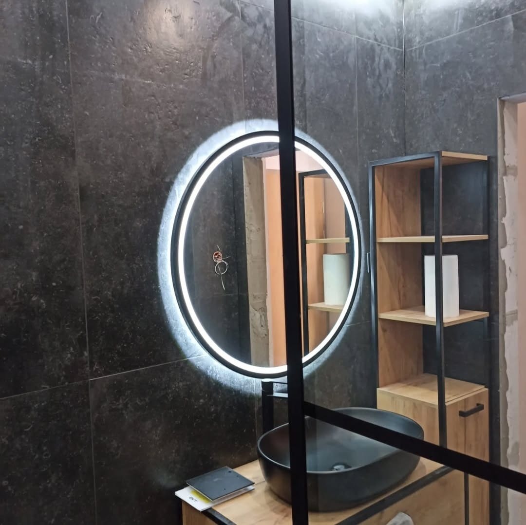 Ciemna łazienka z okrągłym lustrem LED, czarną umywalką nablatową i regałem z drewnianymi półkami, widoczne przewody elektryczne na ścianie.