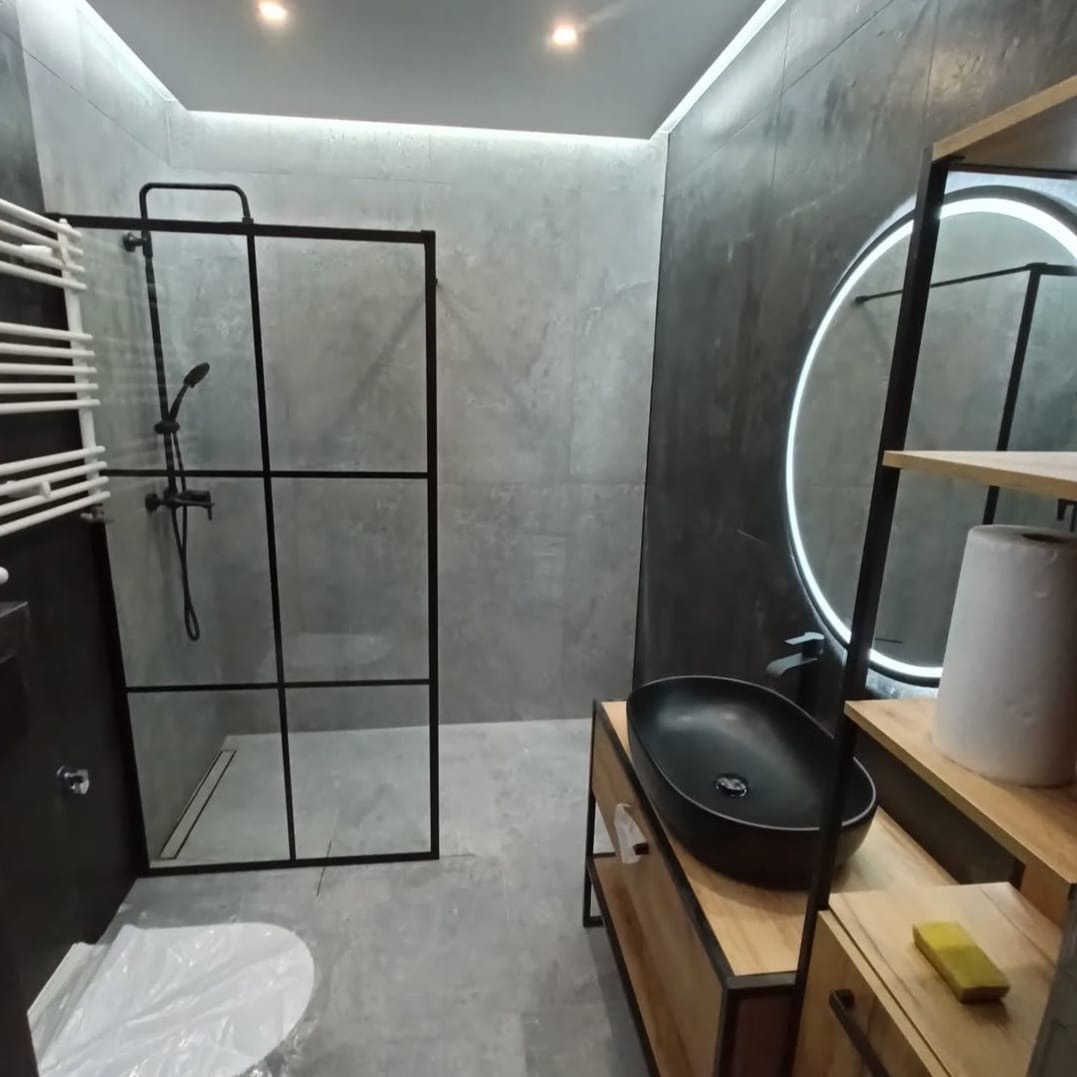 Nowoczesna łazienka z kabiną prysznicową typu walk-in z czarnymi profilami, owalną umywalką nablatową i okrągłym lustrem z podświetleniem LED, wykończona szarą płytką imitującą beton.