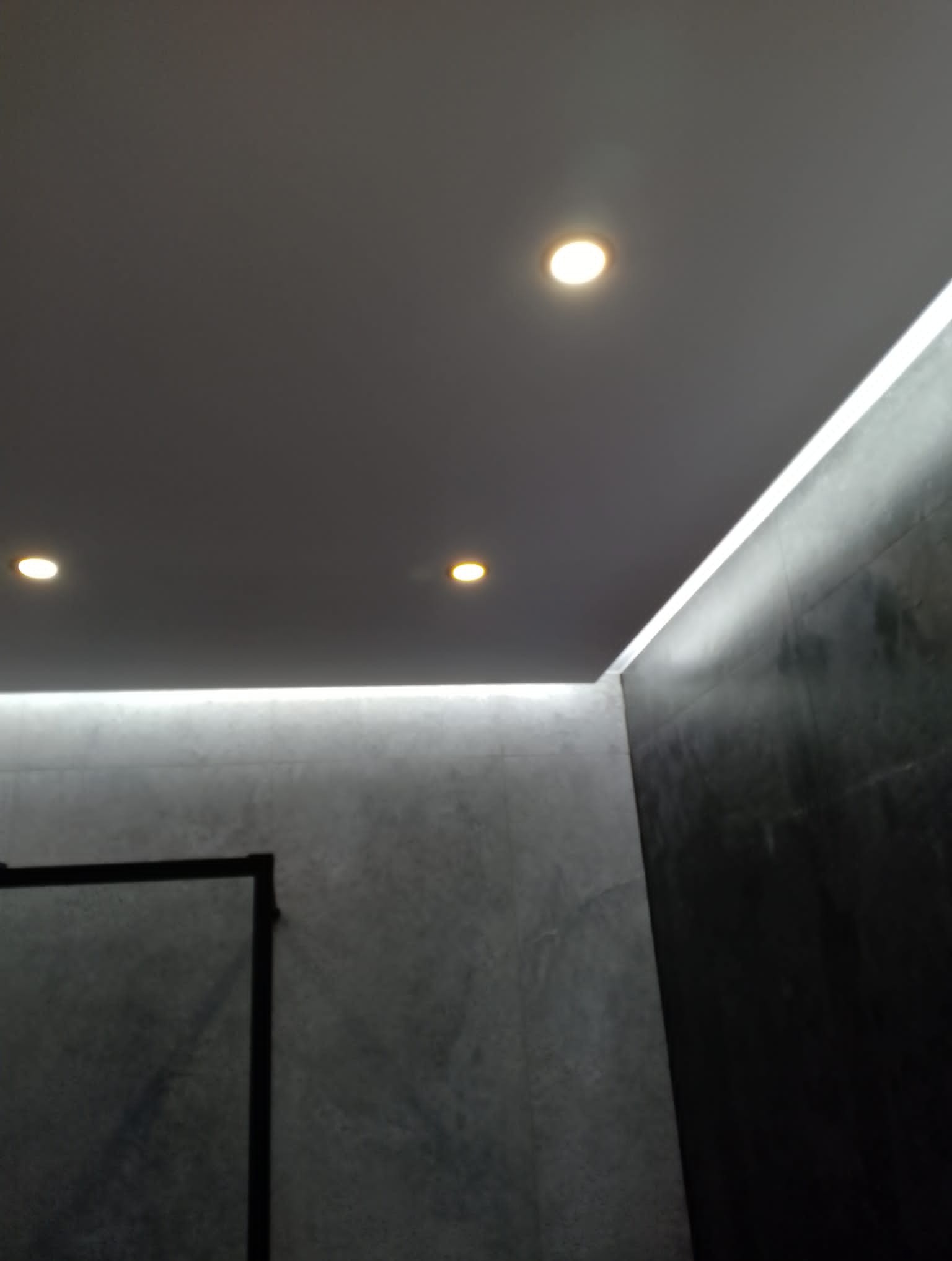 Szare, minimalistyczne wnętrze łazienki z oświetleniem LED podsufitowym i punktowymi lampami, widoczna szklana kabina prysznicowa z czarną ramą.