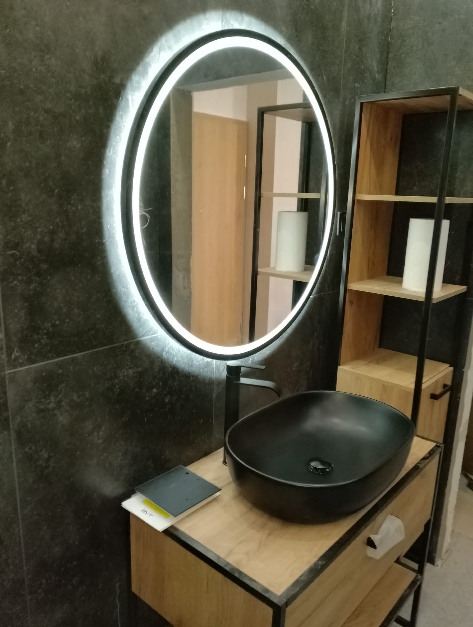 Nowoczesna łazienka z czarną umywalką nablatową, okrągłym lustrem z podświetleniem LED i drewnianym regałem z czarnymi metalowymi ramami, na tle ciemnoszarych płytek.