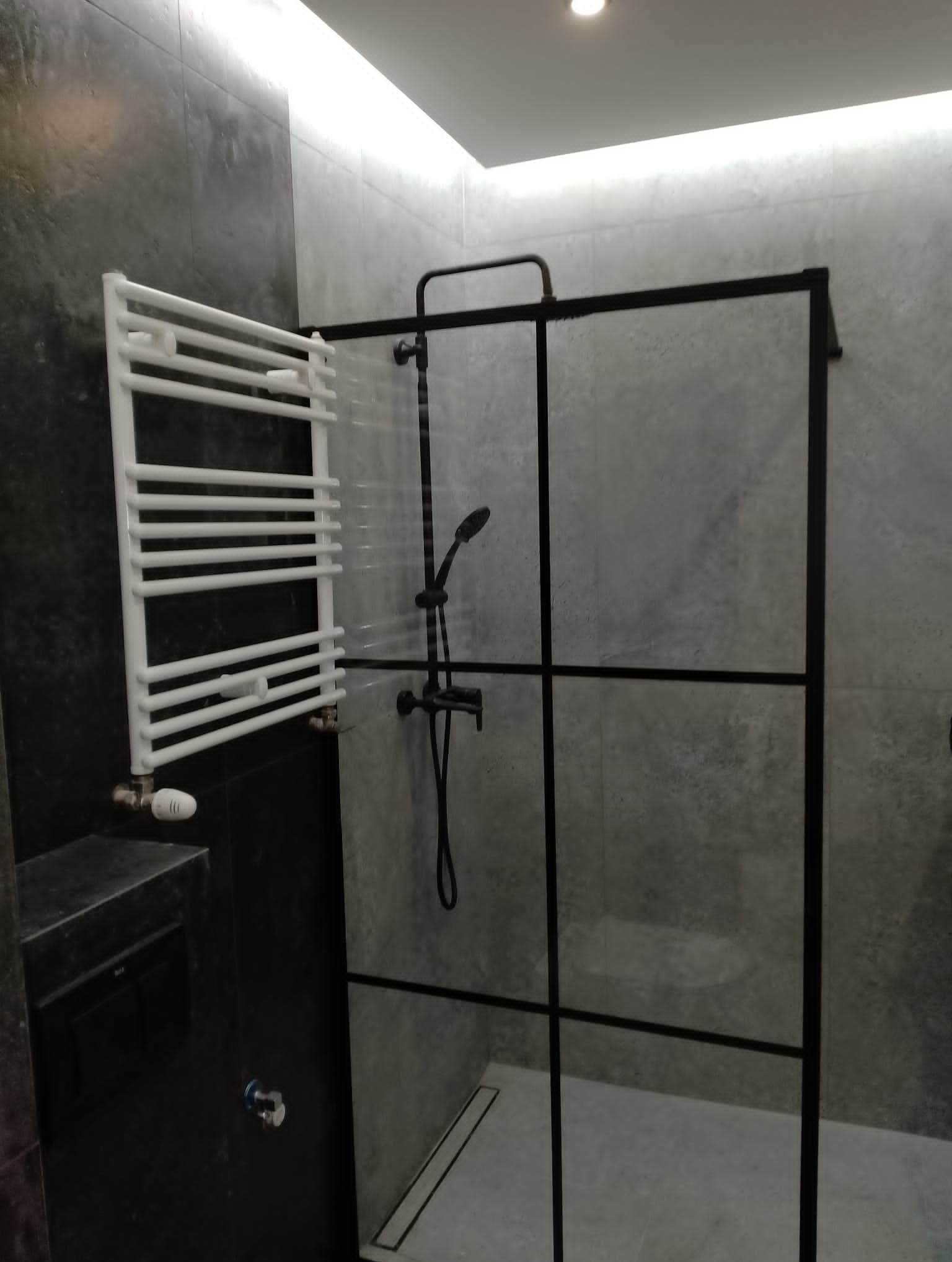 Nowoczesna łazienka z kabiną prysznicową typu walk-in z czarnymi profilami, grzejnikiem drabinkowym w kolorze białym i szarą, teksturowaną ścianą.