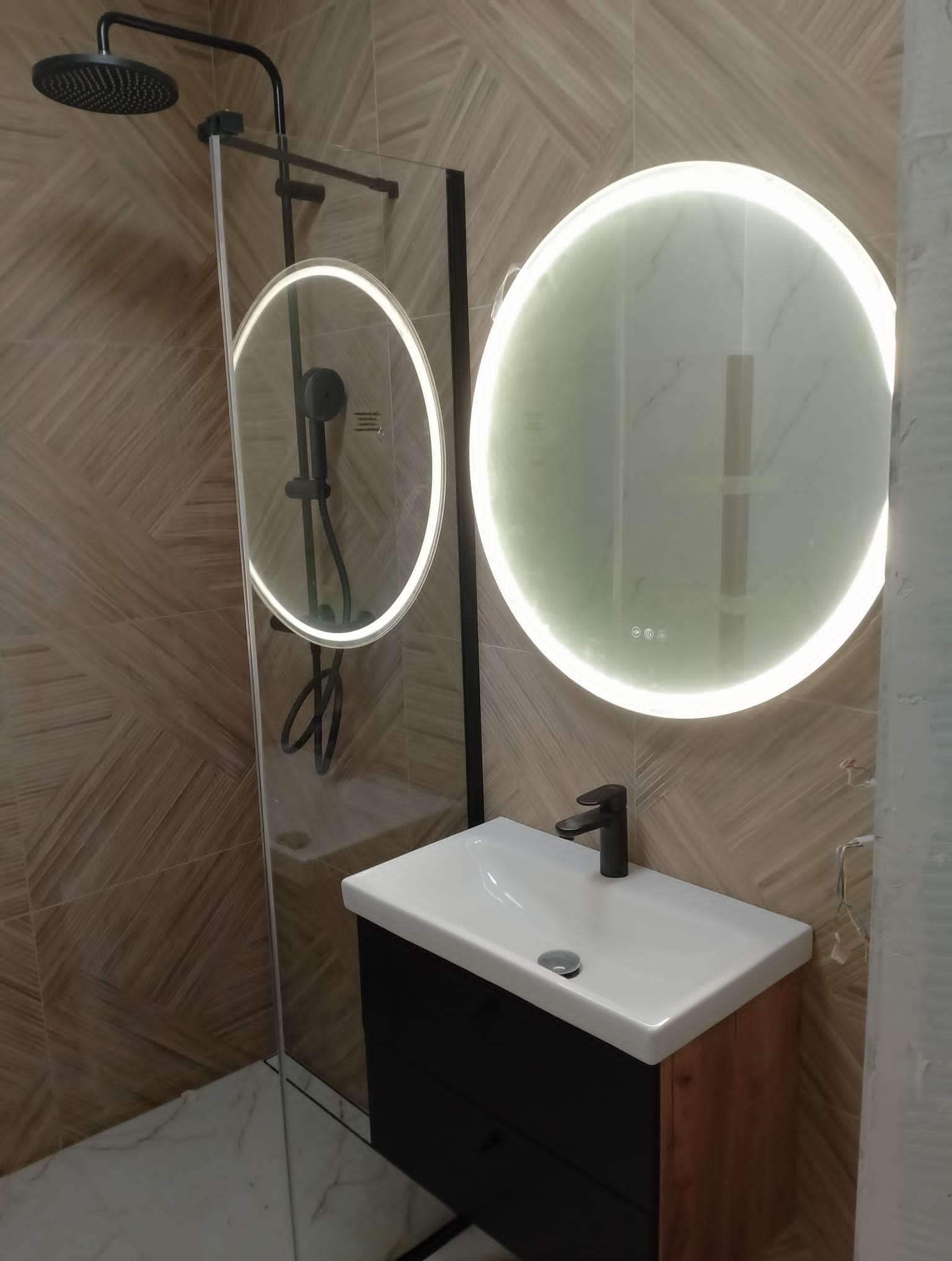 Nowoczesna łazienka z okrągłym lustrem LED, kabiną prysznicową ze szklaną ścianką i umywalką z szafką w kolorze drewna i czerni, na ścianach płytki imitujące drewno ułożone we wzór jodełki.