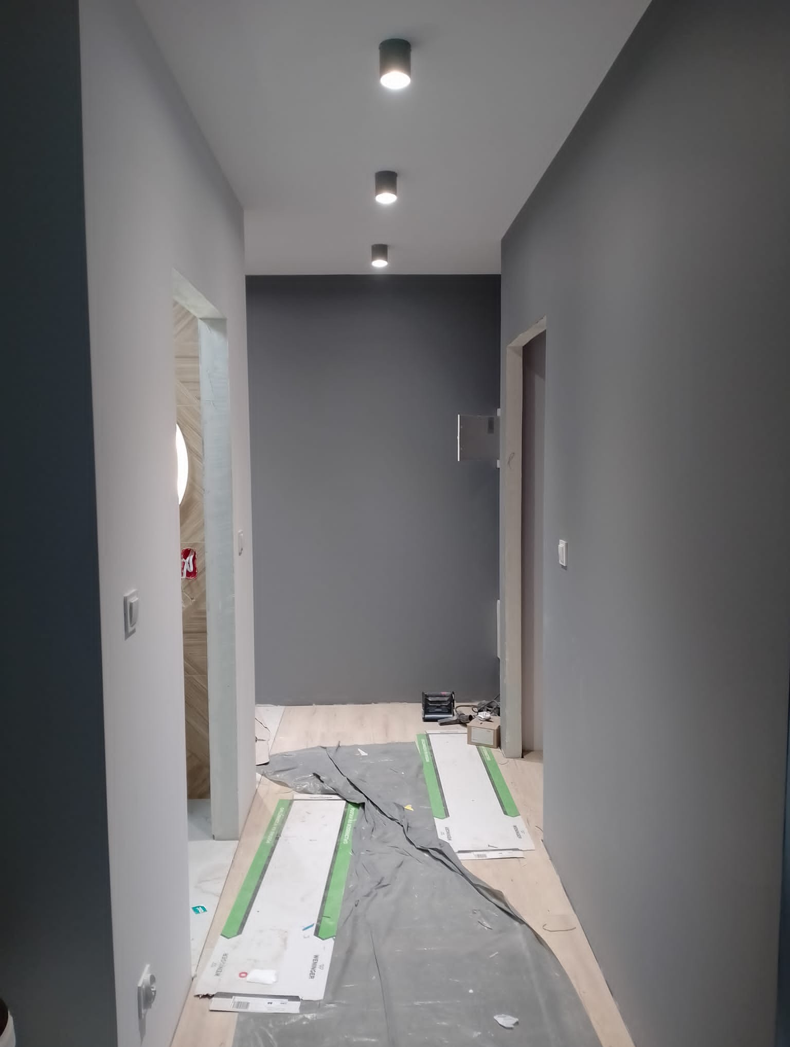 Korytarz w trakcie odświeżania: szare ściany, podłoga zabezpieczona folią malarską, widoczne płyty kartonowo-gipsowe i lampy sufitowe punktowe.