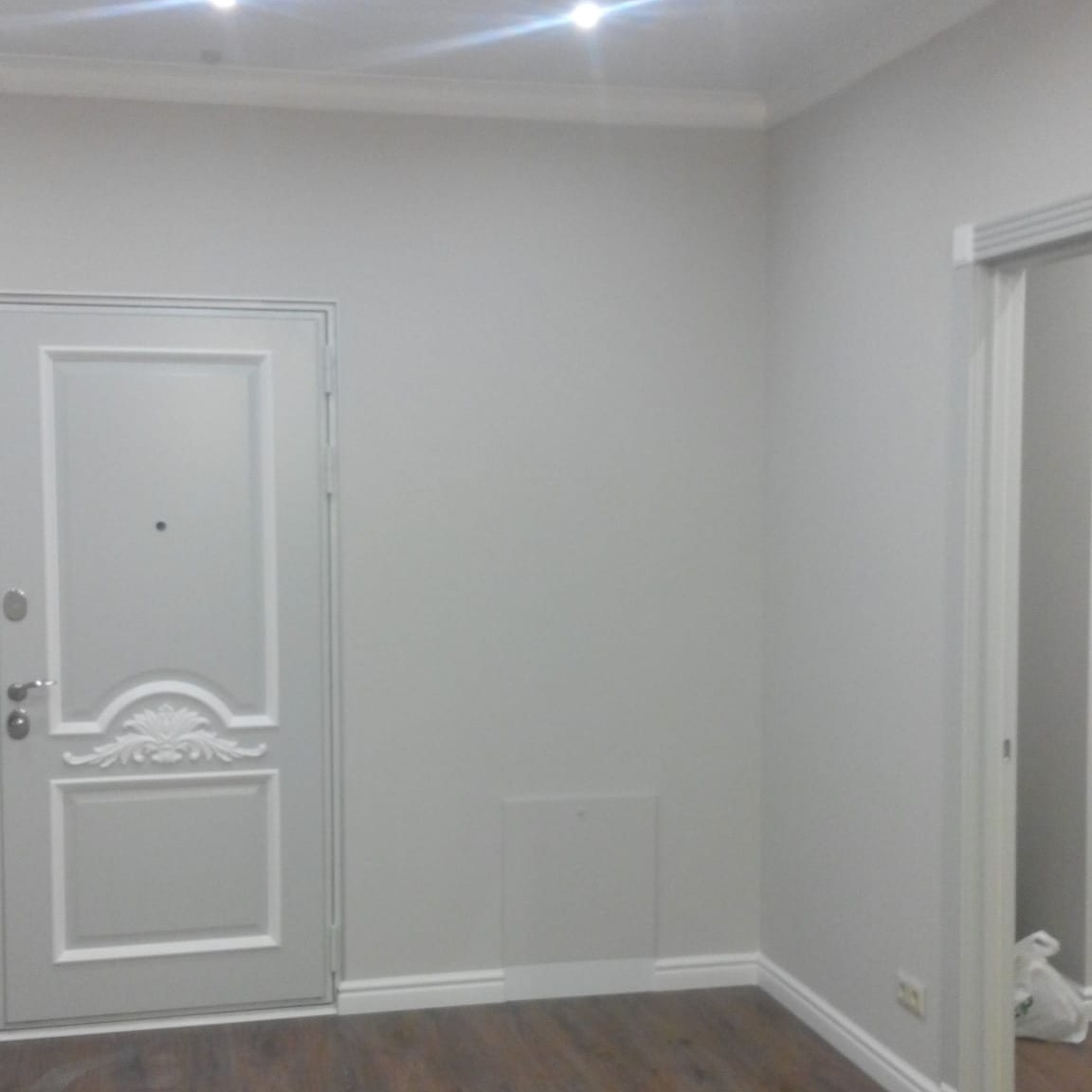 Szare wnętrze po odświeżeniu: widoczne pomalowane ściany, ozdobne listwy przypodłogowe i sufitowe, drewnopodobna podłoga, drzwi wejściowe z dekoracyjnym panelem oraz fragment ościeżnicy drugich drzwi.