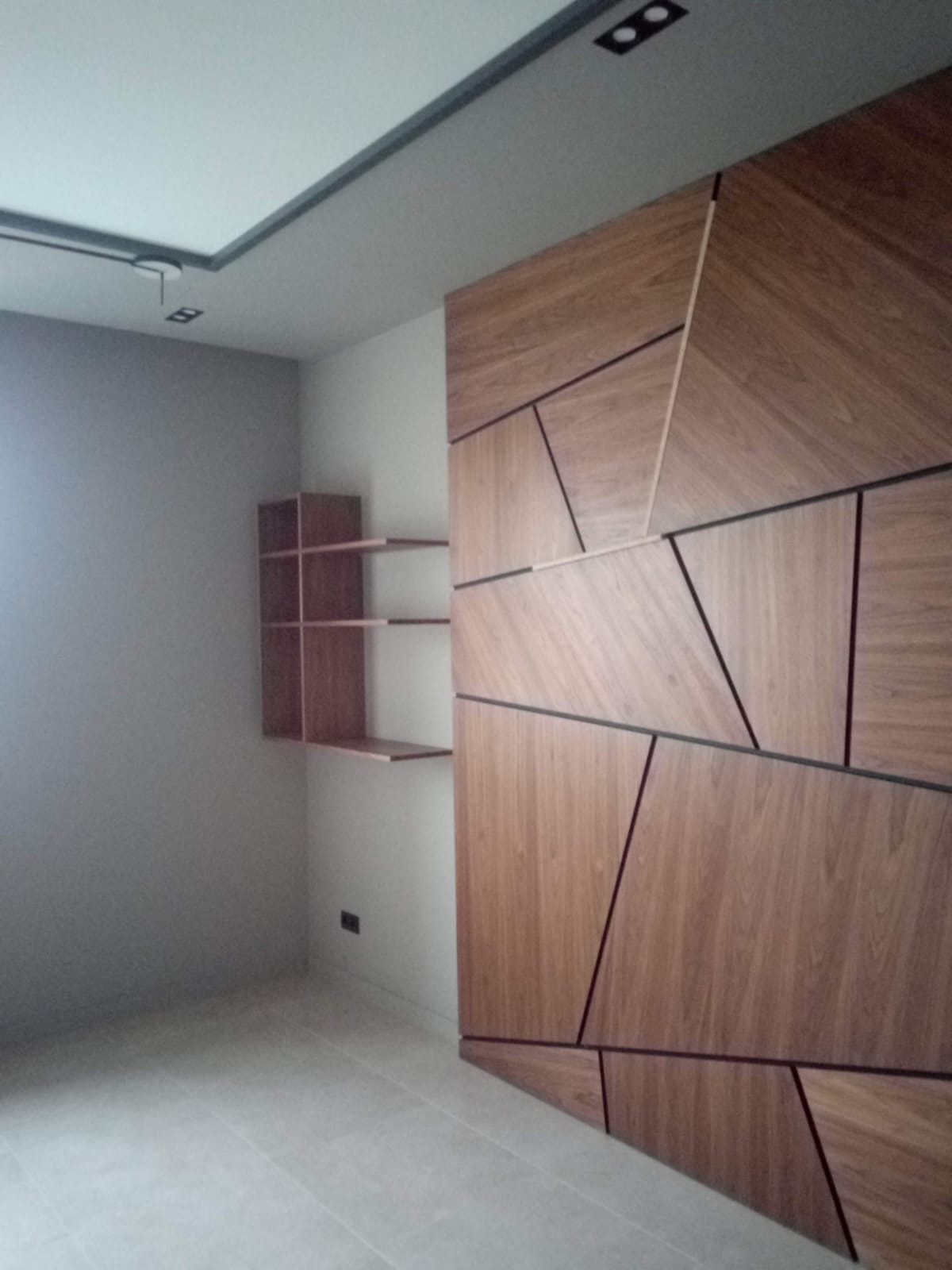 Nowoczesna ściana z paneli drewnianych w geometryczne wzory, minimalistyczne półki wiszące i jasna podłoga w pustym pomieszczeniu.