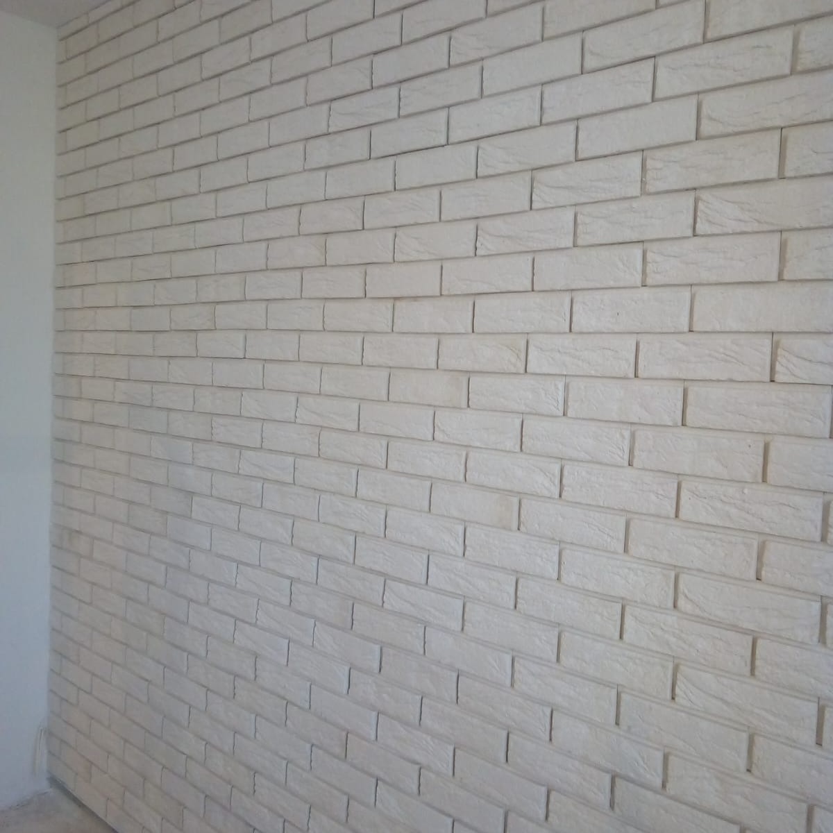 Ściana wyłożona białymi, wąskimi cegłami dekoracyjnymi z delikatną fakturą, widoczne fugi, ujęcie pod kątem z fragmentem białej ściany po lewej stronie.
