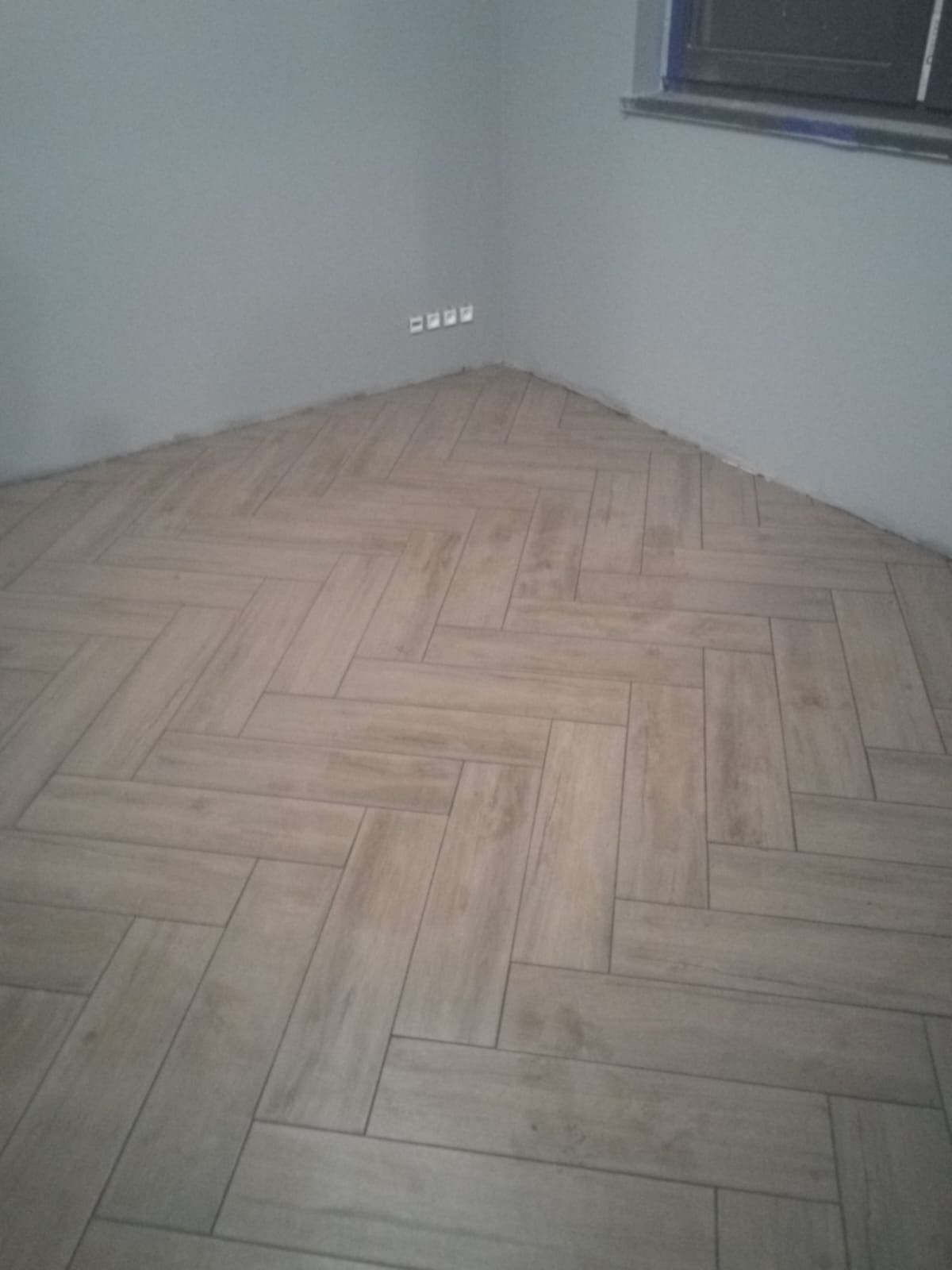 Ułożona w jodełkę podłoga z płytek imitujących drewno w rogu pokoju, widoczne gniazdka elektryczne na szarej ścianie.