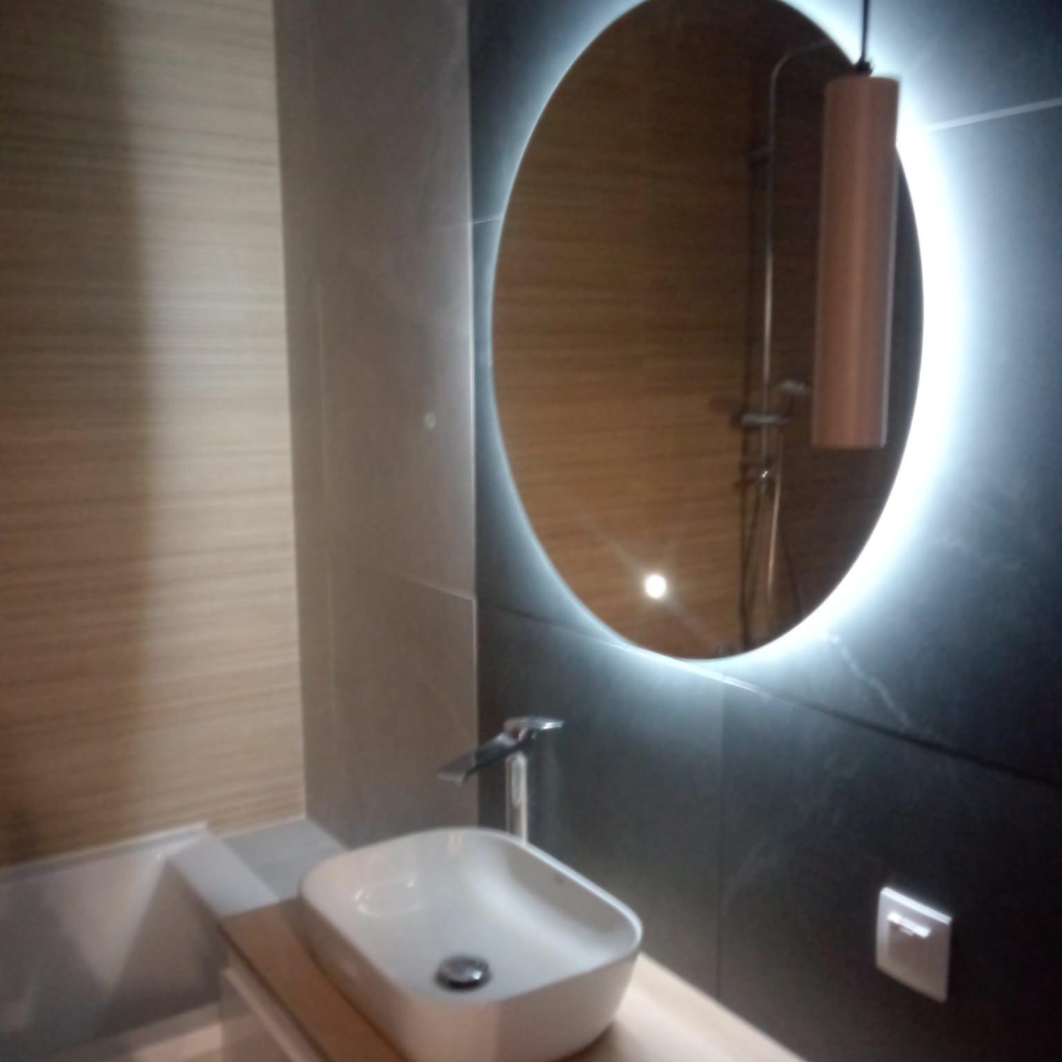 Nowoczesna łazienka z okrągłym lustrem LED, minimalistyczną umywalką nablatową i czarnymi płytkami imitującymi marmur, odbicie kabiny prysznicowej w lustrze.