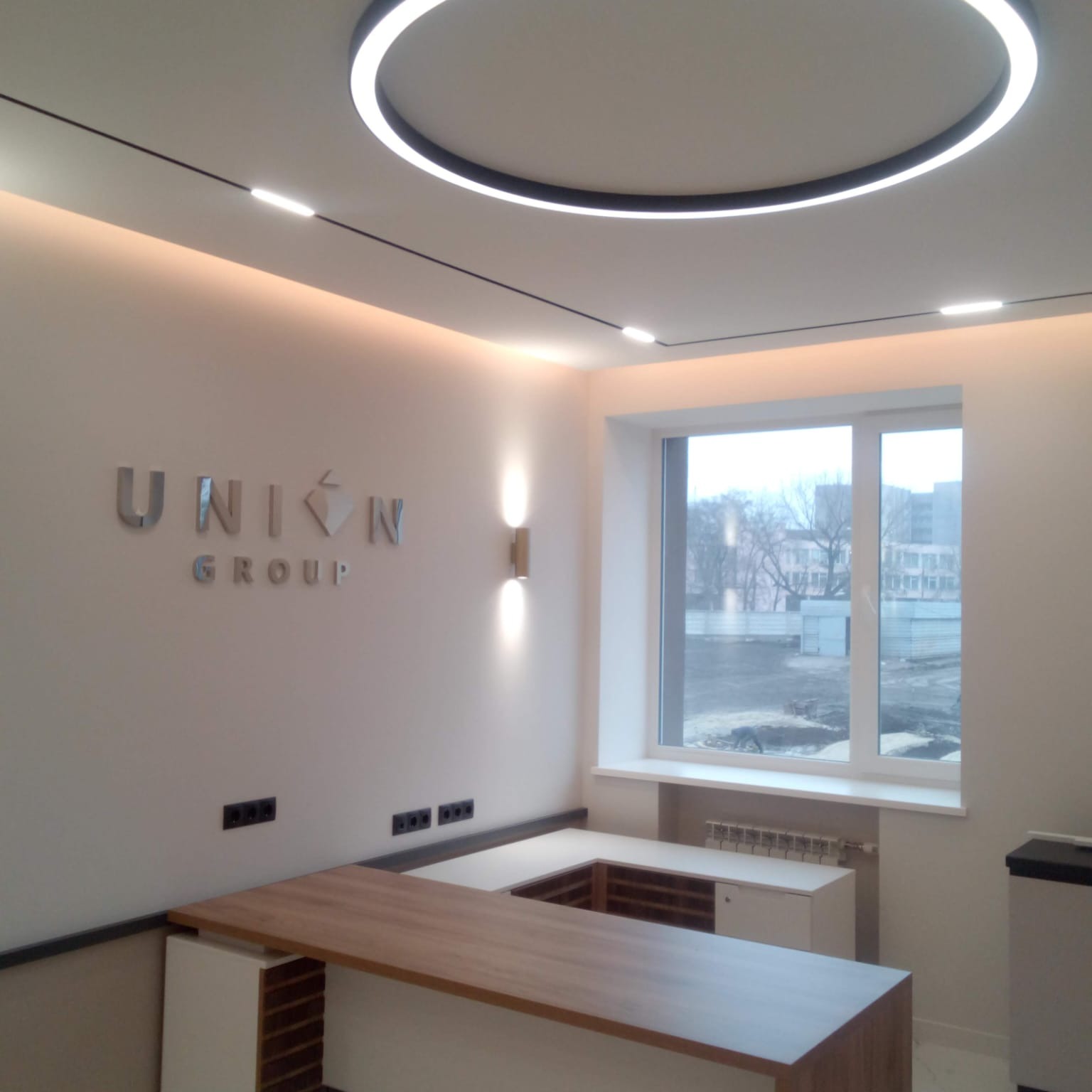 Nowoczesne biuro z oświetleniem LED: okrągła lampa sufitowa, listwy świetlne, logo 'UNION GROUP' na ścianie, drewniane biurko i duże okno z widokiem na plac budowy.