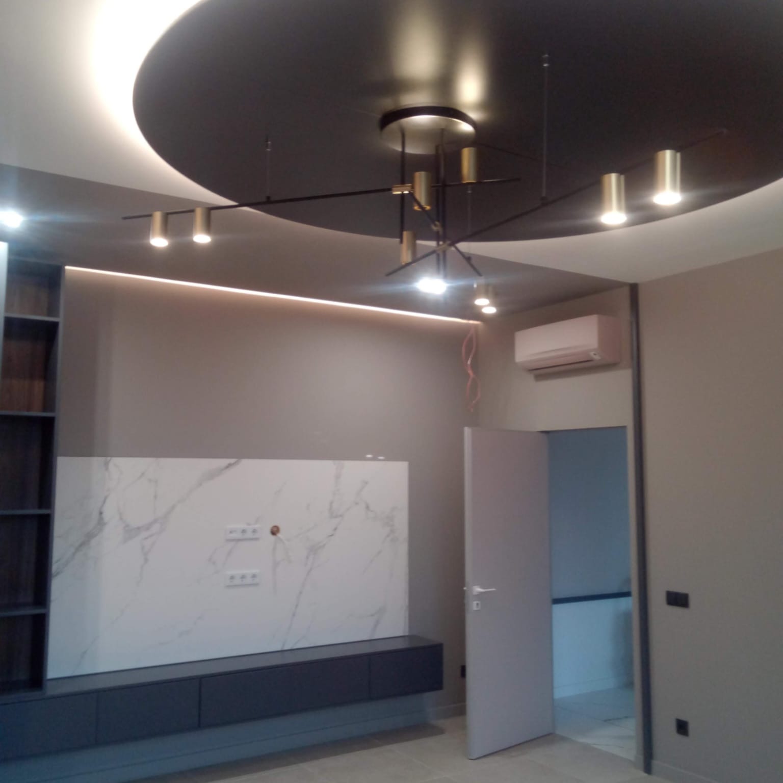 Nowoczesny salon z podwieszanym sufitem, oświetleniem punktowym i designerską lampą wiszącą, ściana wykończona płytami imitującymi marmur, minimalistyczne szafki RTV i klimatyzator.