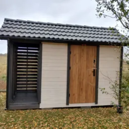 Nowoczesna konstrukcja z drewnianą wstawką i zadaszeniem z blachodachówki, otoczona opadłymi liśćmi, z otwartą przestrzenią po lewej stronie.