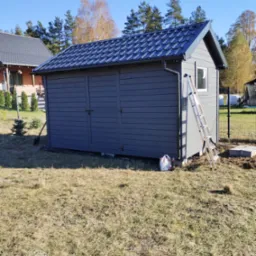 Szara drewniana altana z dwuspadowym dachem z blachy, drabina oparta o ścianę, widoczne drzewa w tle i fragment sąsiedniego domu.