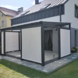 Nowoczesna konstrukcja z czarnych belek i białych paneli zlokalizowana przy domu z szarym dachem, otoczona trawnikiem i kostką brukową.