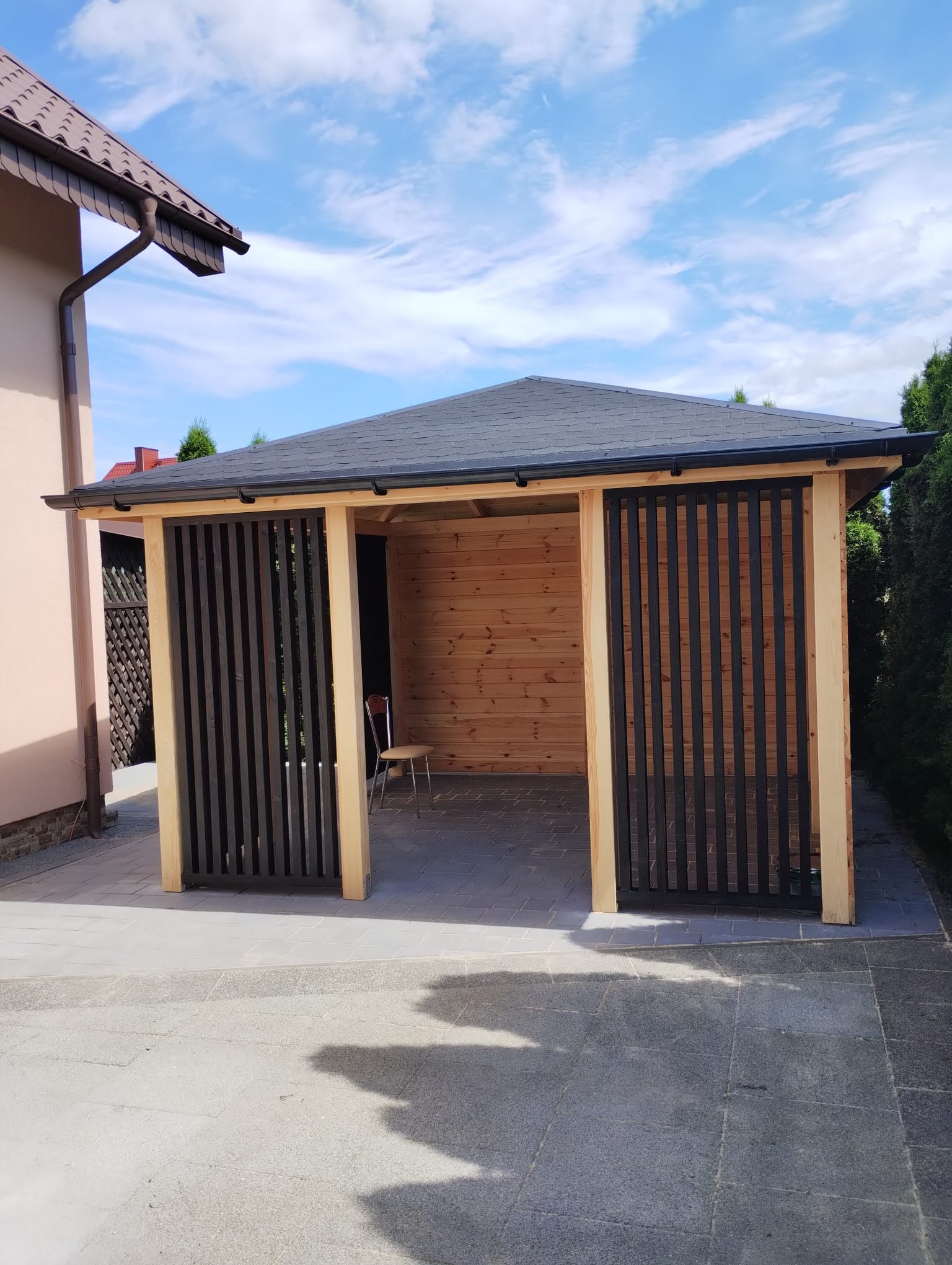 Drewniana konstrukcja z czarnym dachem i pionowymi, ciemnymi panelami, częściowo zacieniona, na tle błękitnego nieba z chmurami.
