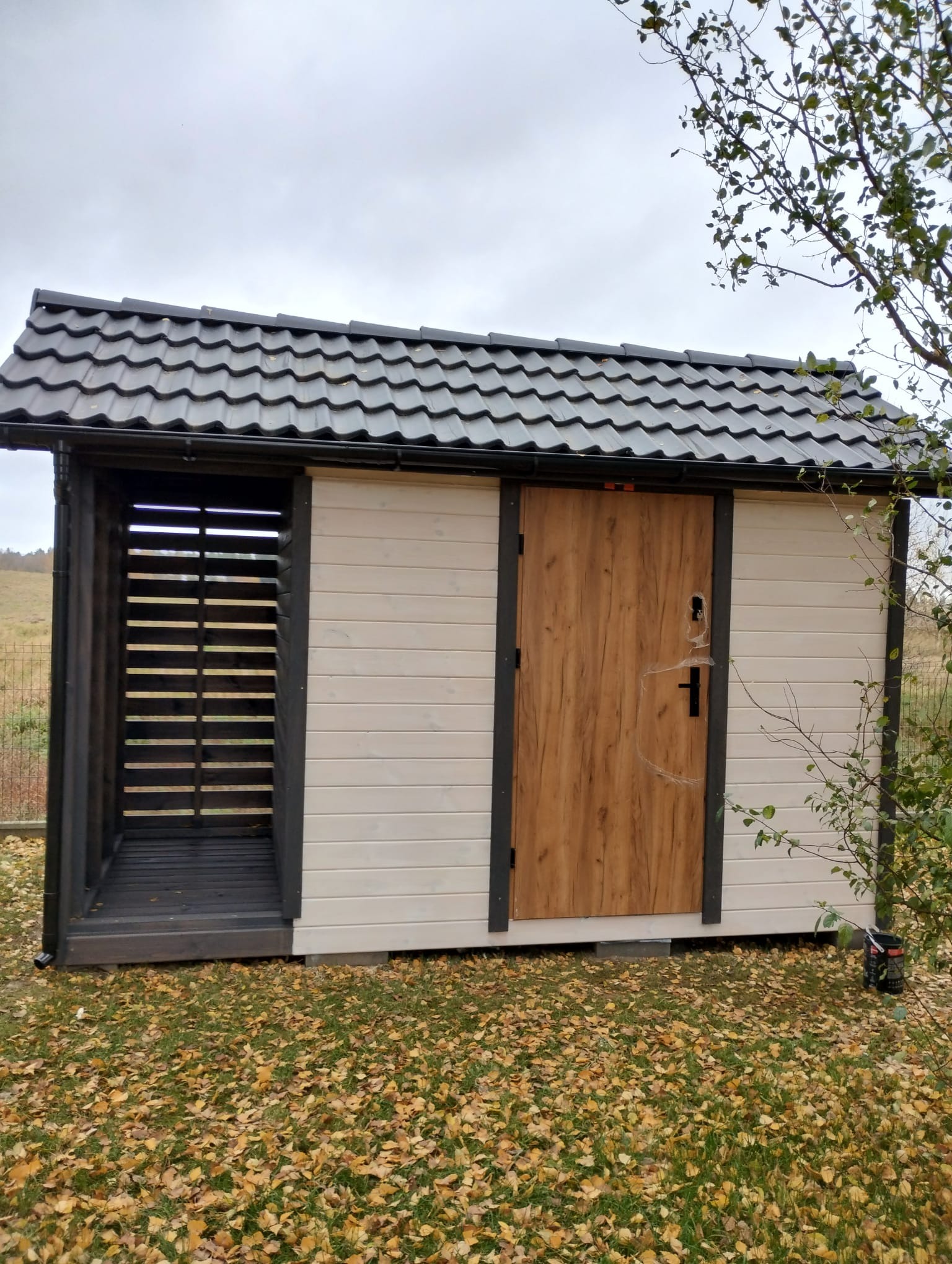 Nowoczesna konstrukcja z drewnianą wstawką i zadaszeniem z blachodachówki, otoczona opadłymi liśćmi, z otwartą przestrzenią po lewej stronie.