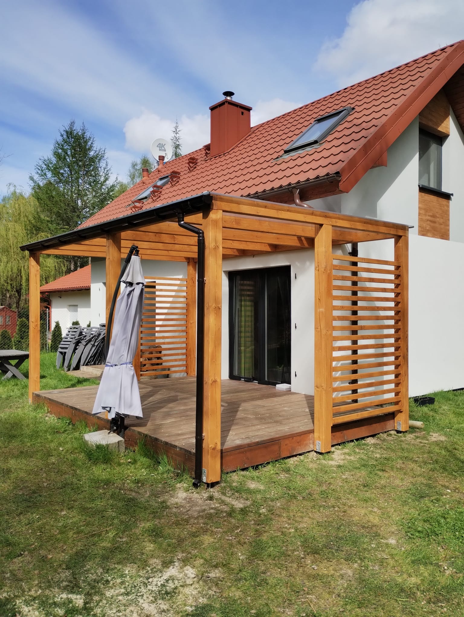 Drewniana konstrukcja z zadaszeniem i ażurowymi ściankami, przylegająca do budynku z czerwonym dachem. Na tarasie złożony parasol przeciwsłoneczny. Widoczne rynny odprowadzające wodę z dachu.