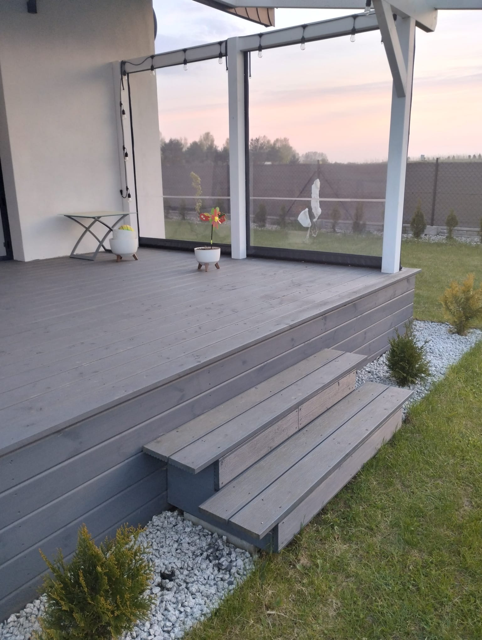 Szare deski tarasowe z trzema stopniami, widok na ogród z elementami dekoracyjnymi, takimi jak doniczki z kwiatami i lampki girlandy.