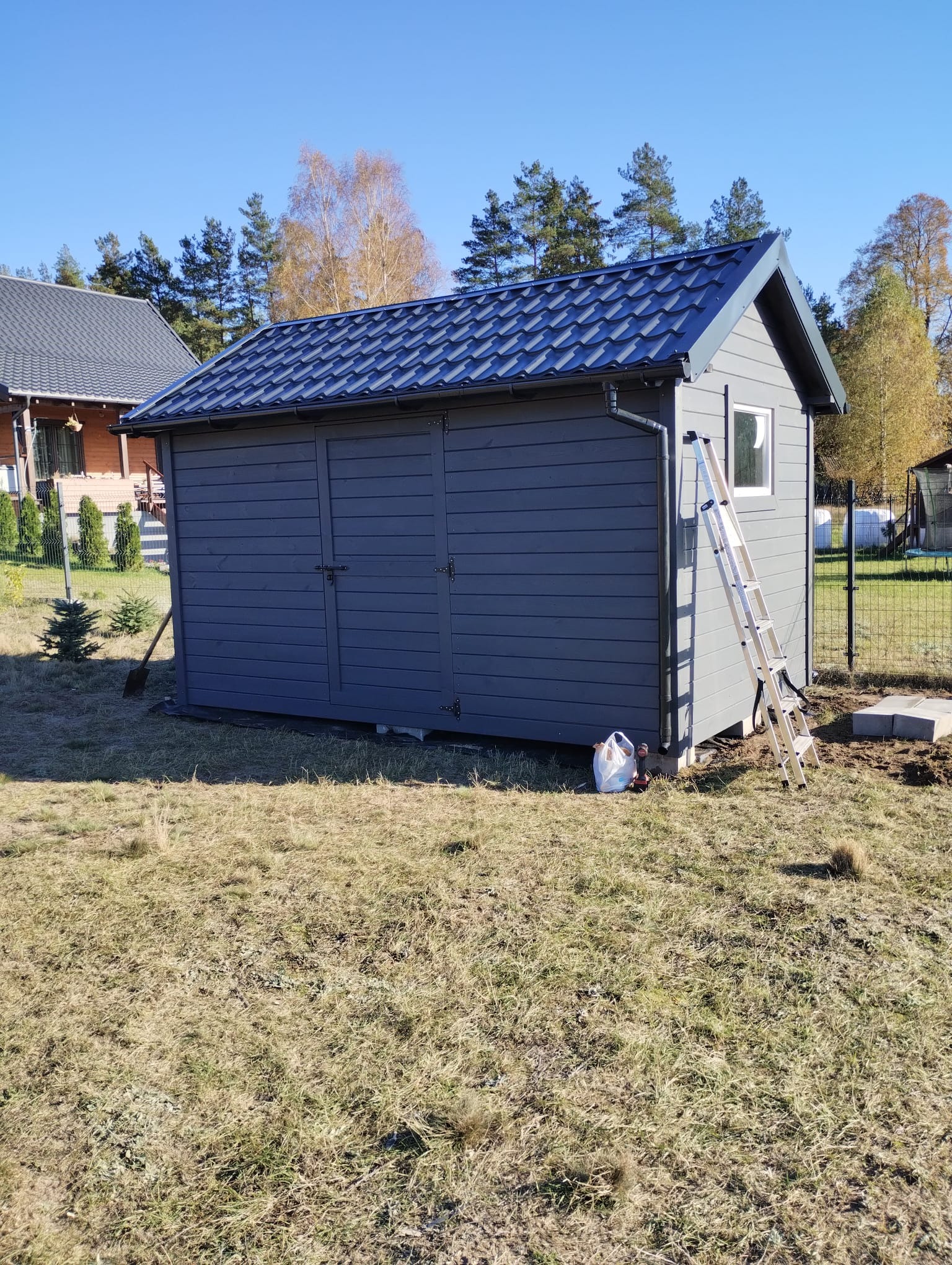 Szara drewniana altana z dwuspadowym dachem z blachy, drabina oparta o ścianę, widoczne drzewa w tle i fragment sąsiedniego domu.