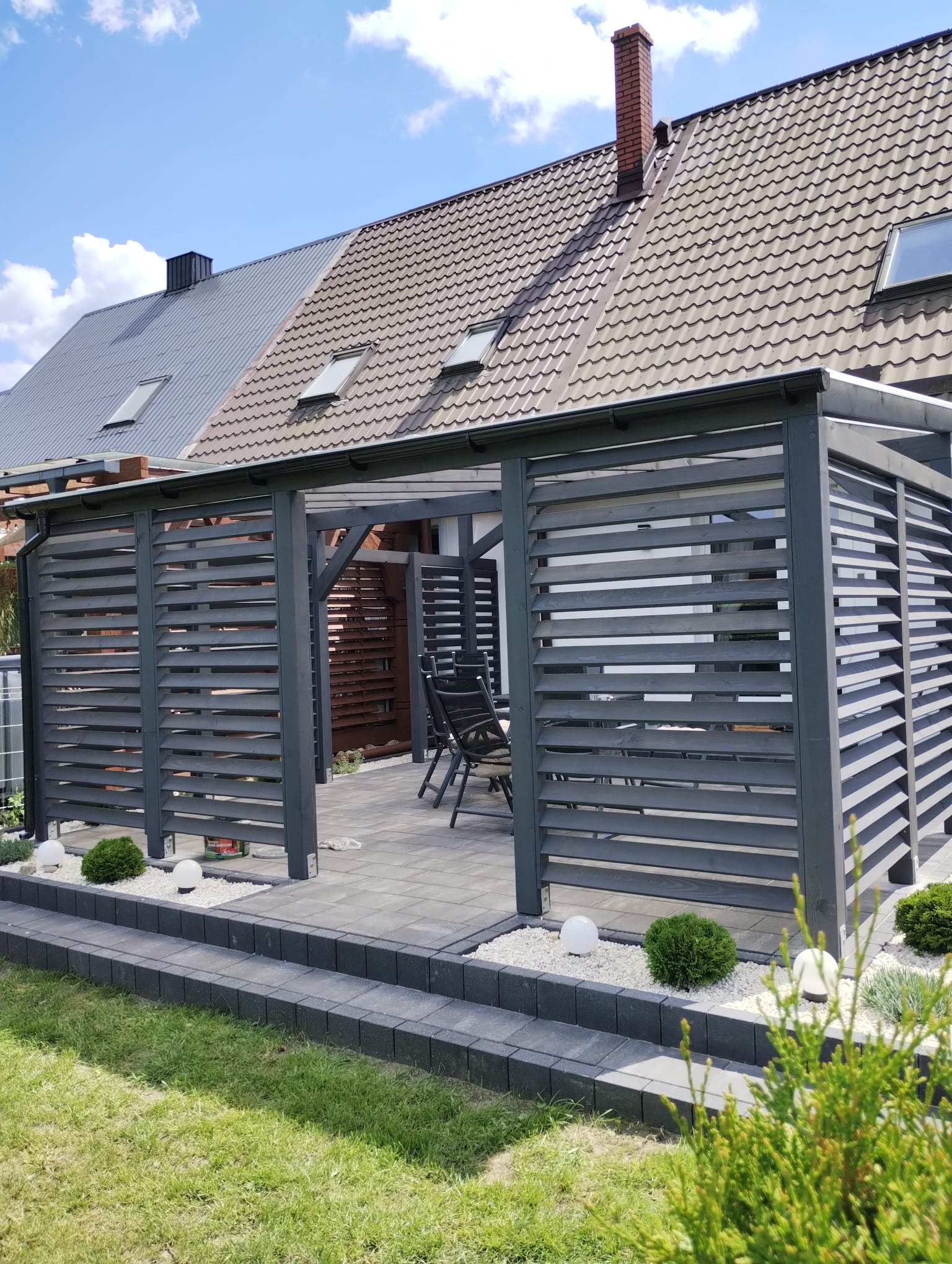 Nowoczesna, szara konstrukcja z poziomych desek, tworząca zacienioną przestrzeń z meblami ogrodowymi na wyłożonym kostką tarasie, z niskimi krzewami i lampami kulami w białym żwirze, na tle domu...