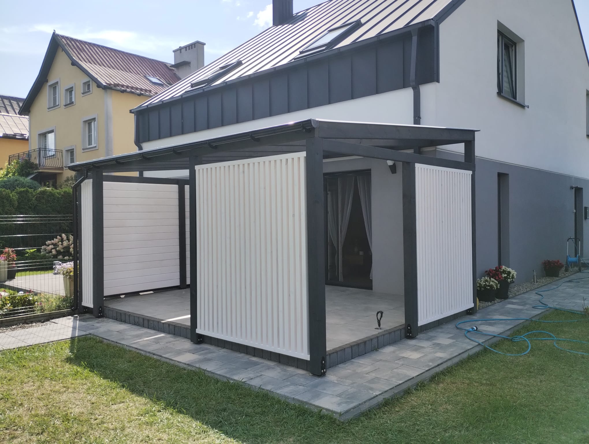 Nowoczesna konstrukcja z czarnych belek i białych paneli zlokalizowana przy domu z szarym dachem, otoczona trawnikiem i kostką brukową.