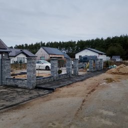 Brukarstwo i ogrody - Nowoczesne ogrodzenie z szarej kostki łupanej w trakcie montażu, widoczne słupki i podmurówka, w tle domy jednorodzinne z panelami słonecznymi i sterta piasku.