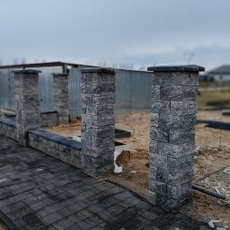 Brukarstwo i ogrody - Fragment budowanego ogrodzenia z szarej kostki, widoczny fragment utwardzonej kostką drogi, w tle blaszany garaż i dom w budowie.