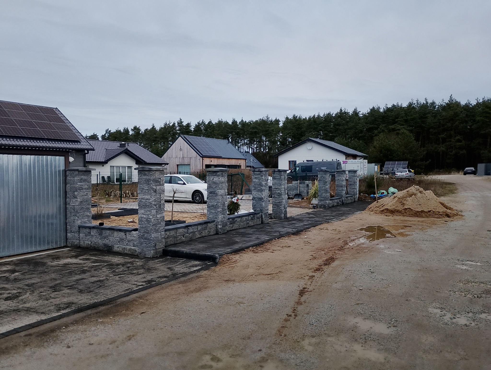 Nowoczesne ogrodzenie z szarej kostki łupanej w trakcie montażu, widoczne słupki i podmurówka, w tle domy jednorodzinne z panelami słonecznymi i sterta piasku.