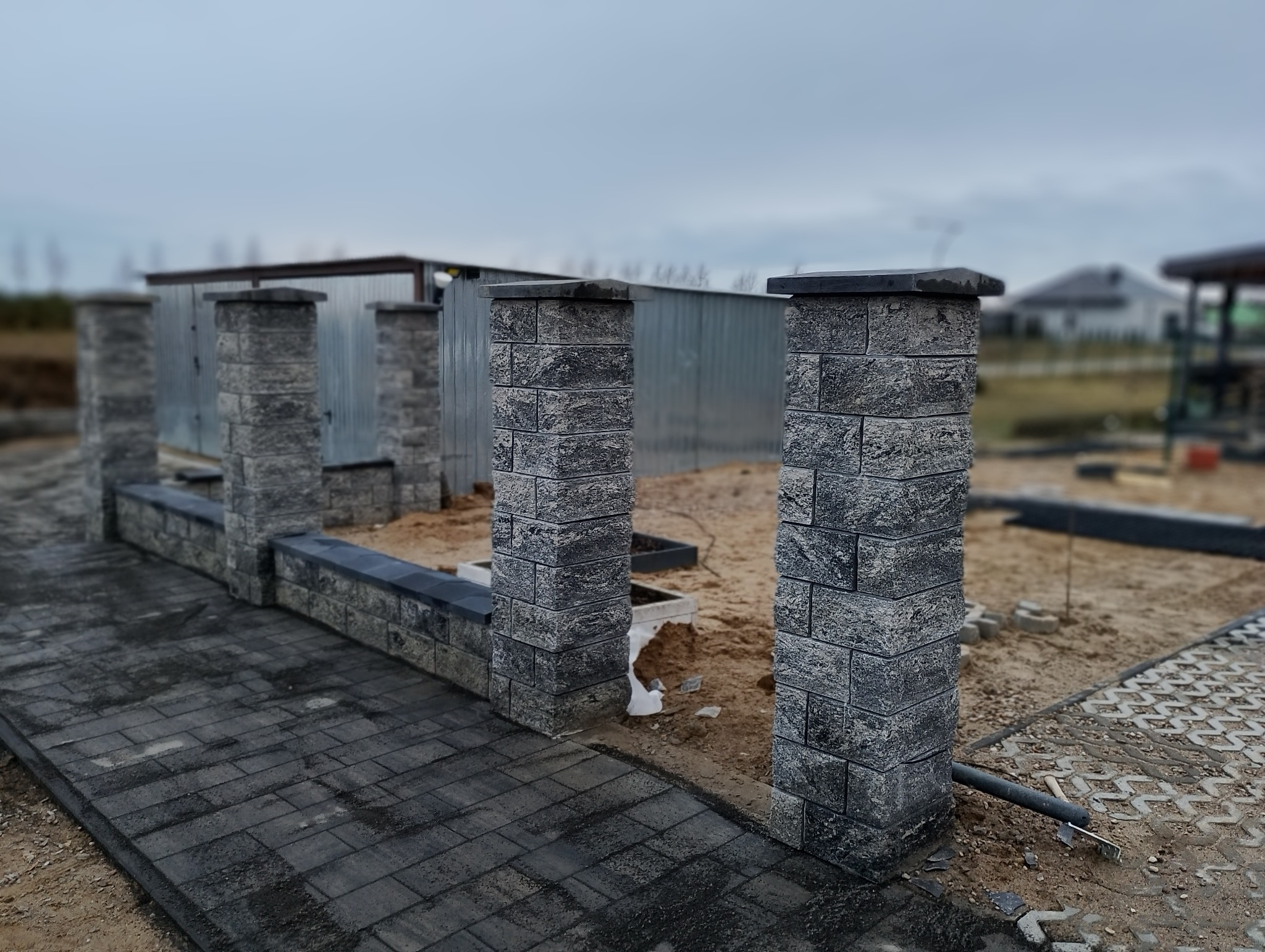 Fragment budowanego ogrodzenia z szarej kostki, widoczny fragment utwardzonej kostką drogi, w tle blaszany garaż i dom w budowie.