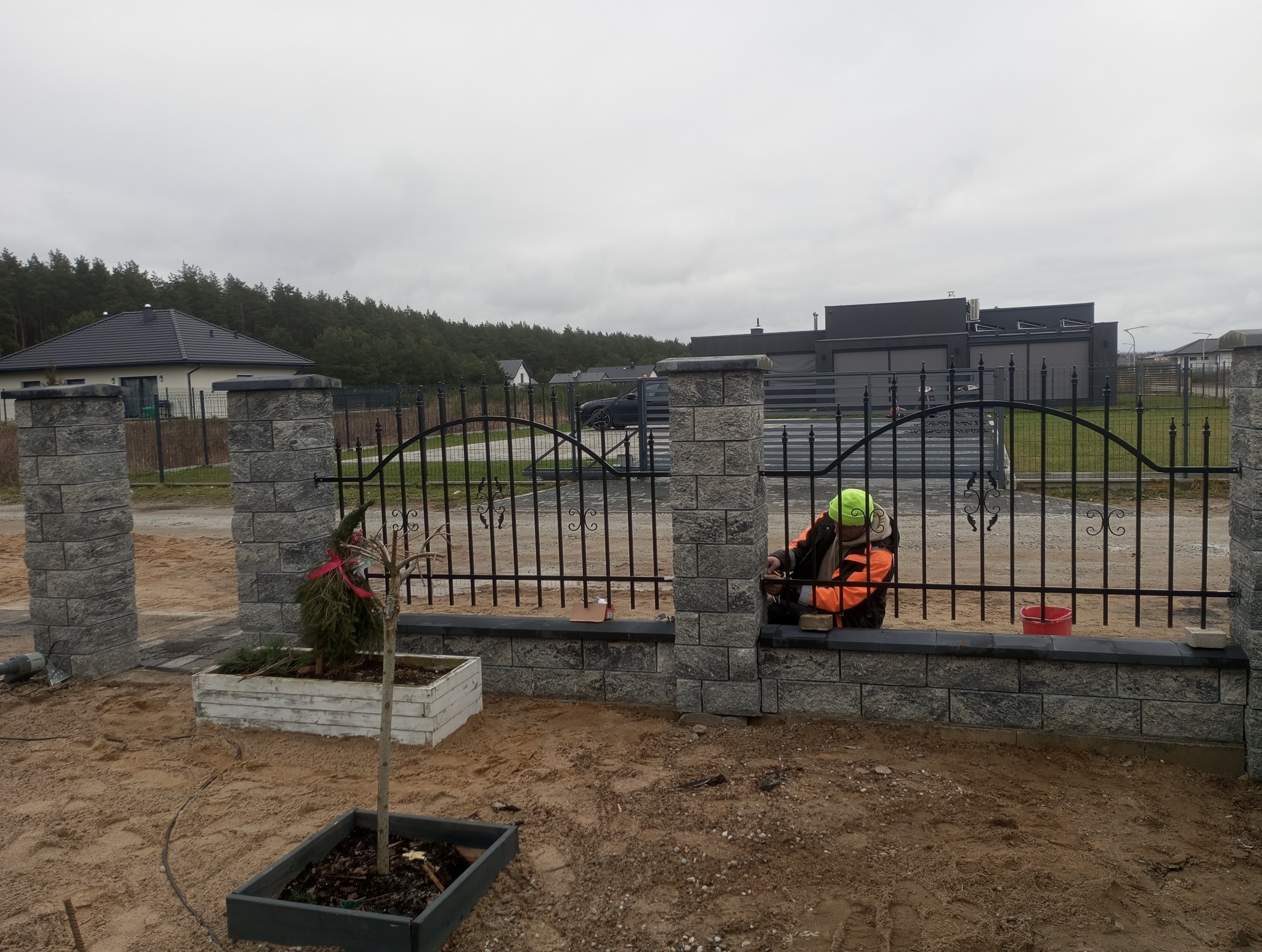 Montaż ozdobnego ogrodzenia z kamiennymi słupkami i kutymi przęsłami w Gryficach, pracownik w odblaskowej kamizelce podczas prac wykończeniowych, widoczne drzewka w donicach.