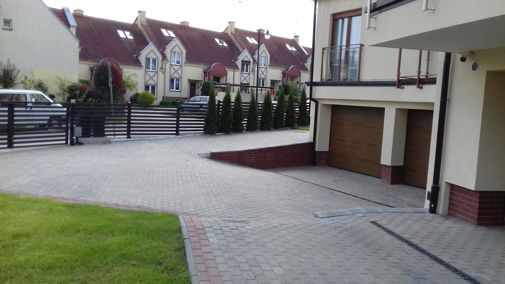 Podjazd z kostki brukowej przed nowoczesnym domem z dwoma garażami, widoczny fragment trawnika i czarne ogrodzenie panelowe.