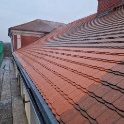 Czyściuch- mycie ciśnieniowe - Częściowo umyty dach z czerwonej dachówki karpiówki, widoczna różnica w kolorze i czystości, rynna oraz komin z cegły w tle, pochmurne niebo.