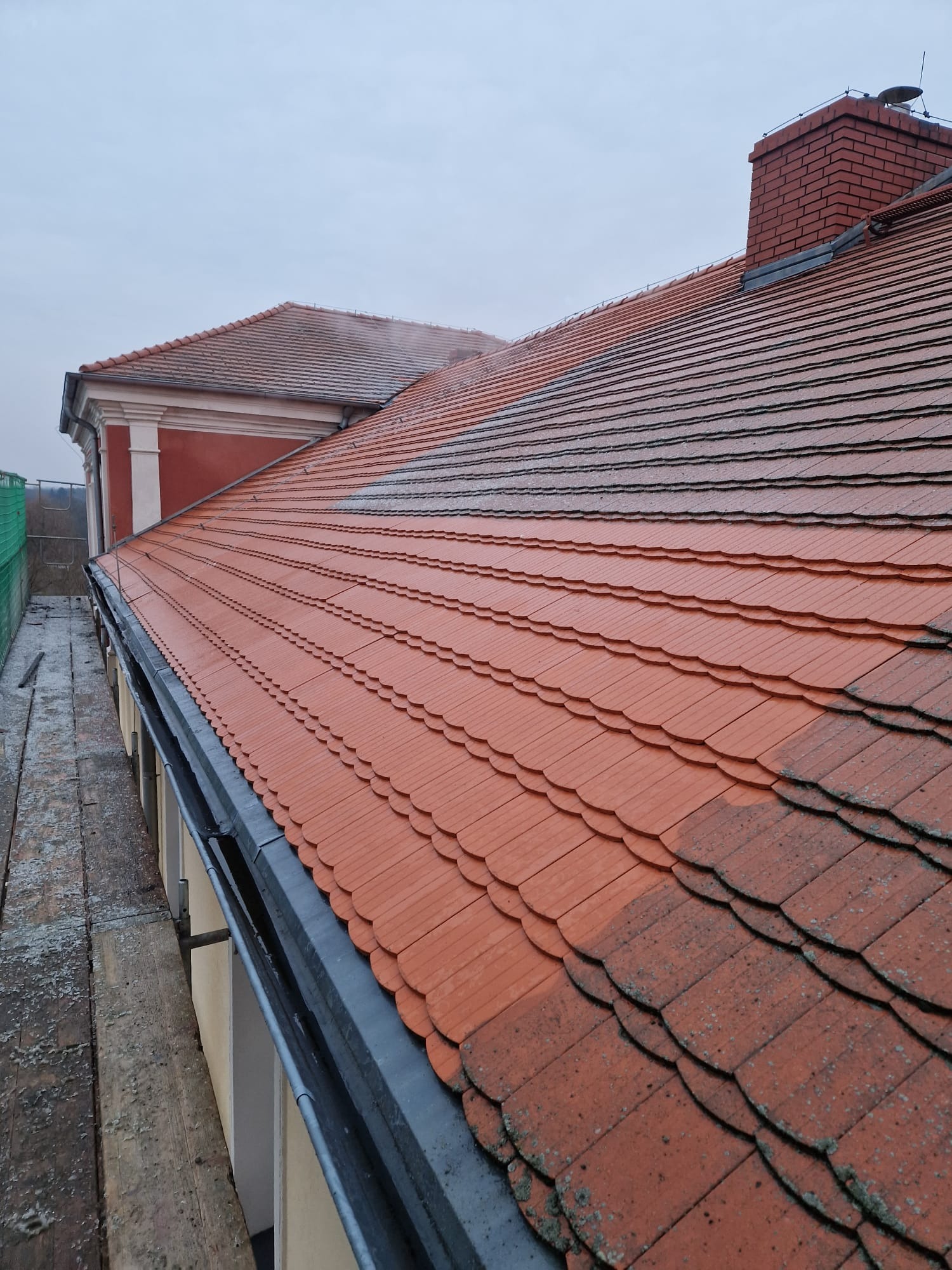Częściowo umyty dach z czerwonej dachówki karpiówki, widoczna różnica w kolorze i czystości, rynna oraz komin z cegły w tle, pochmurne niebo.