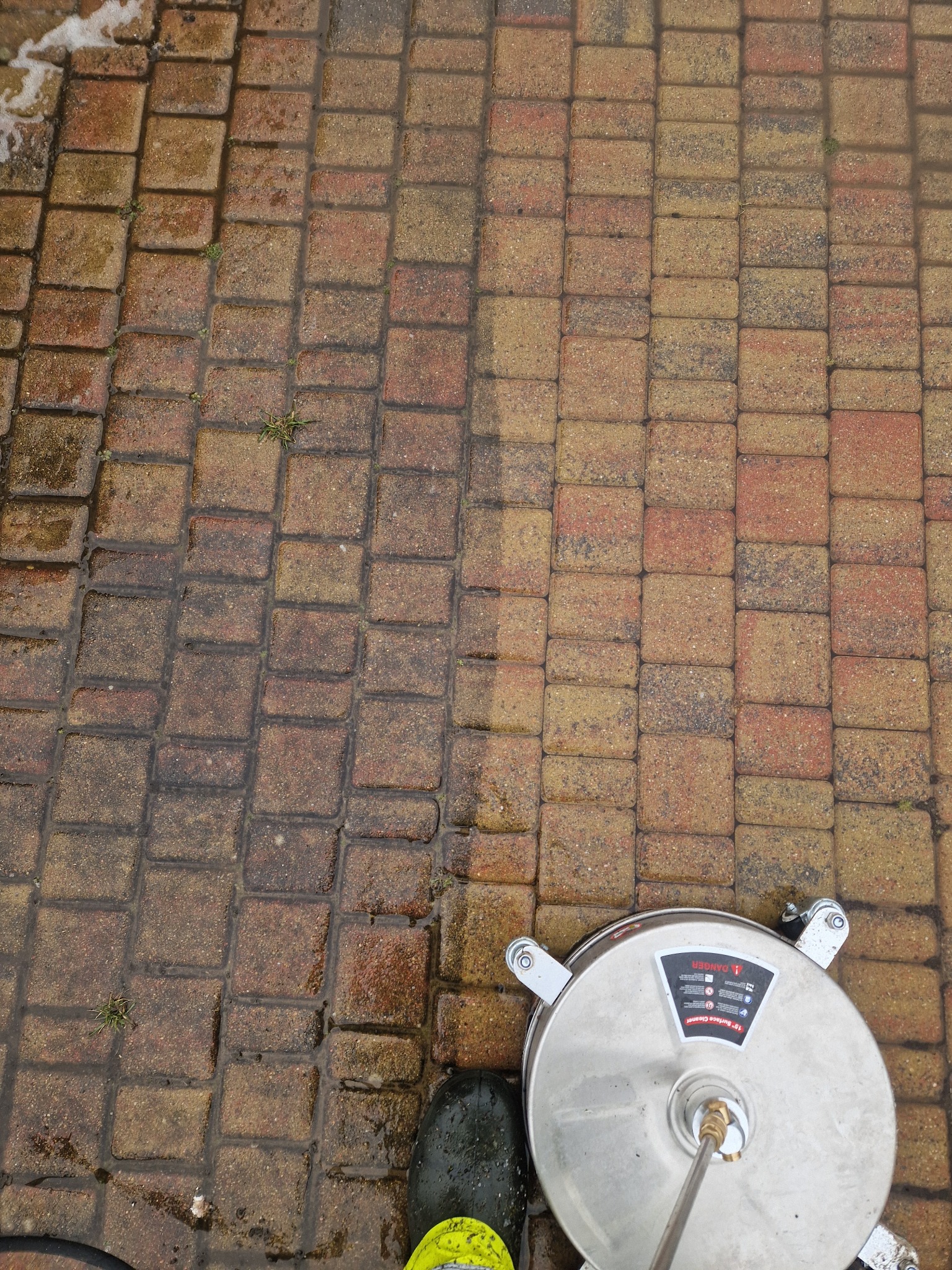 Mycie kostki brukowej z użyciem okrągłej myjki ciśnieniowej; widoczna różnica w kolorze kostki przed i po czyszczeniu, fragment buta operatora w jaskrawym kolorze.