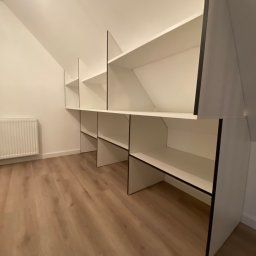 Da Vinci Sztuka Mebli - Białe półki z czarnymi obramowaniami wkomponowane w skos poddasza, widoczny grzejnik i panele podłogowe.