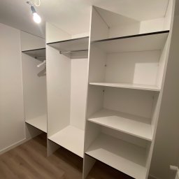 Da Vinci Sztuka Mebli - Wnętrze garderoby z białymi półkami i drążkami na ubrania, widoczna żarówka bez klosza, drewniana podłoga.