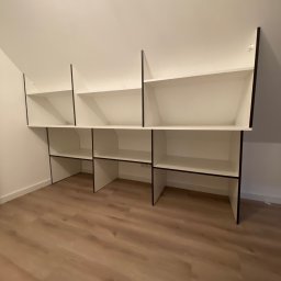 Da Vinci Sztuka Mebli - Białe półki z czarnymi obramowaniami, zamontowane pod skosem dachu, w pustym pomieszczeniu z panelami podłogowymi.