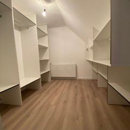 Da Vinci Sztuka Mebli - Pusta garderoba w skosach poddasza, białe półki i wieszaki zamontowane na ścianach, widoczny grzejnik i drewniana podłoga.