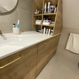 Da Vinci Sztuka Mebli - Łazienka z drewnianą szafką pod umywalką z szufladami i otwartymi półkami, na których ustawione są kosmetyki i akcesoria do makijażu, okrągłe lustro nad umywalką, ręcznik na wieszaku z boku.