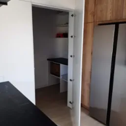 Nowoczesna kuchnia z otwartą szafą wnękową, widoczne półki z artykułami spożywczymi i pudełkami, obok stoi duża lodówka.