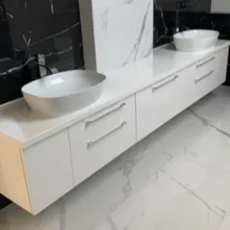 Biała, podwieszana szafka łazienkowa z dwiema owalnymi umywalkami na blacie, minimalistyczne uchwyty, tło z czarno-białych marmurowych płytek, połyskująca podłoga.