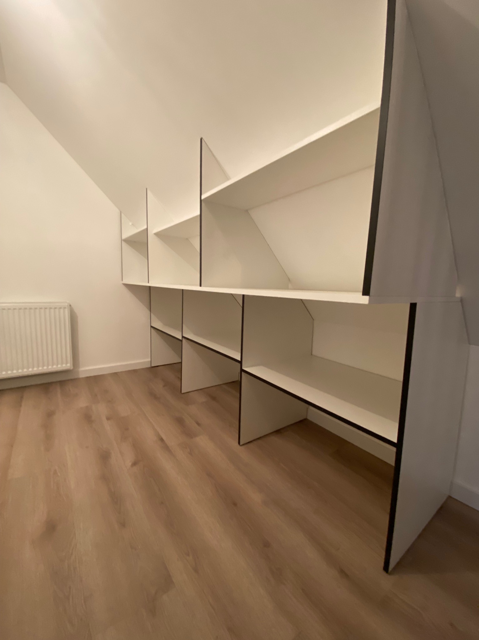 Białe półki z czarnymi obramowaniami wkomponowane w skos poddasza, widoczny grzejnik i panele podłogowe.